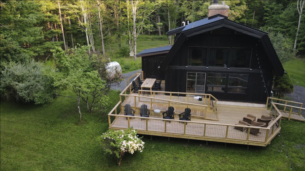 The cabin&rsquo;s front deck with Adirondack chairs, an outdoor dining table, and the barrel sauna at the tree line