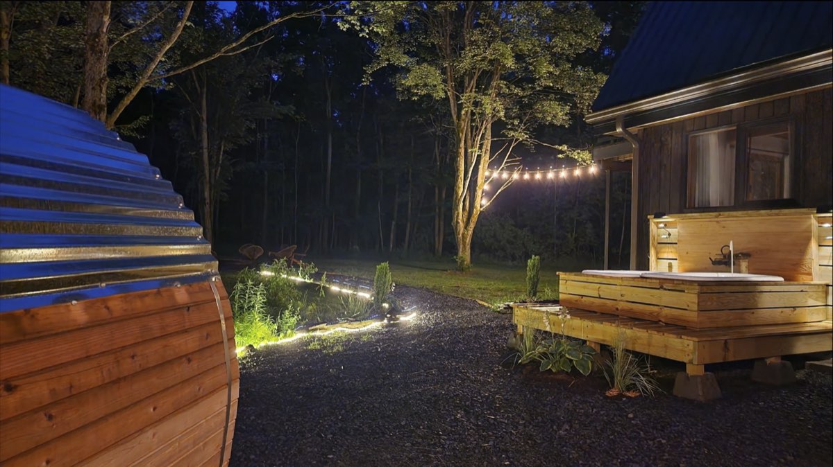 Night view past the barrel sauna to the soaking-tub deck, with string lights in the trees