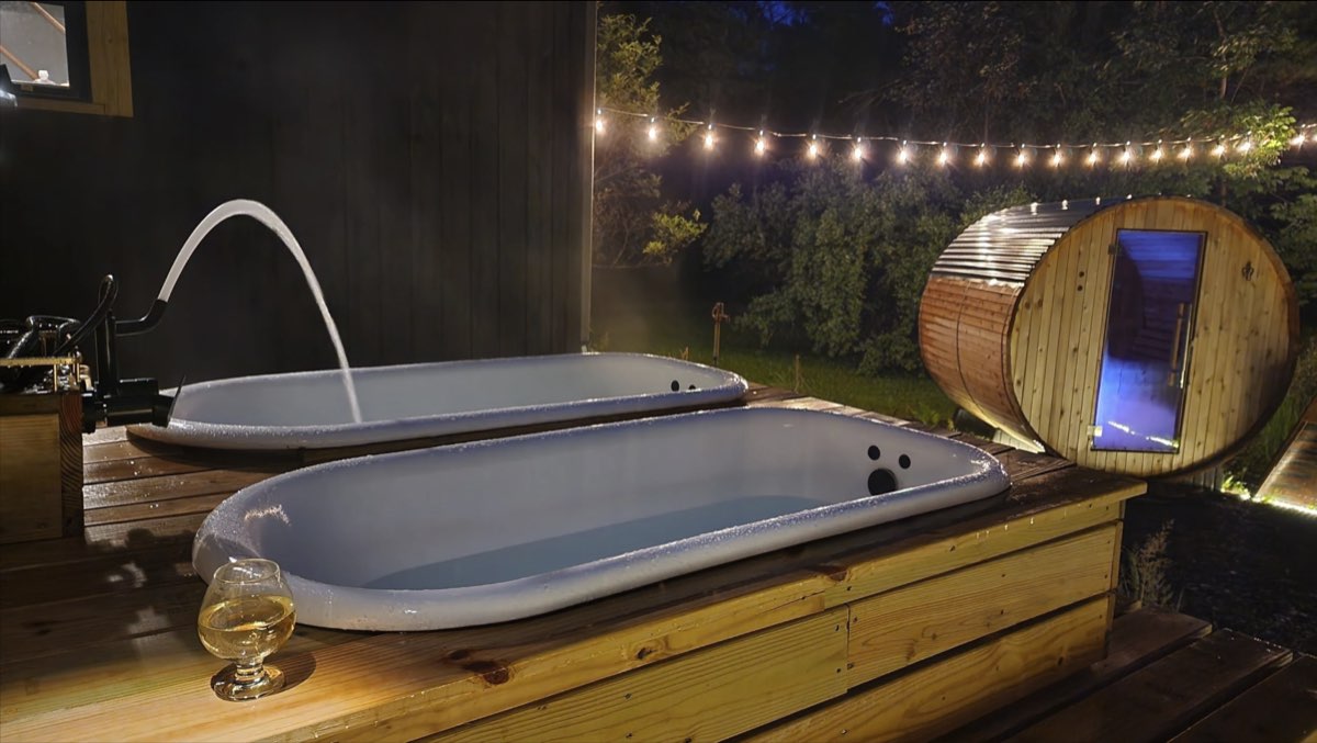 Two outdoor soaking tubs on a wooden deck at night, one filling from a spout, with the lit barrel sauna behind
